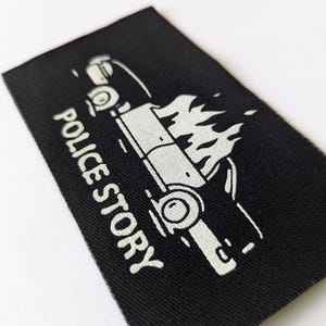 May include: A black fabric patch with a white outline of a car on fire and the text "POLICE STORY" underneath.