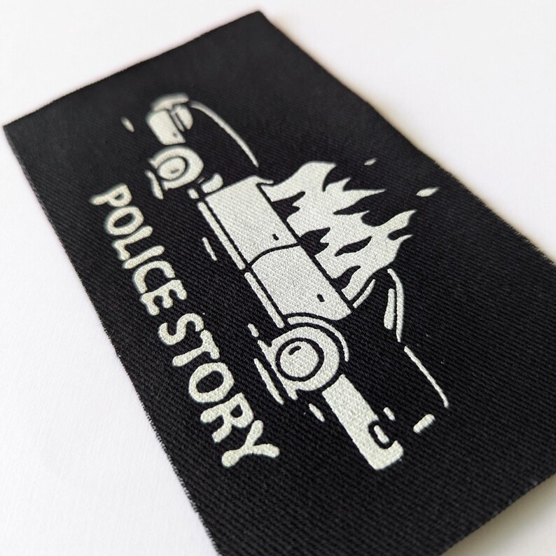 May include: A black fabric patch with a white outline of a car on fire and the text "POLICE STORY" underneath.