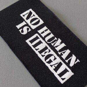 May include: Black rectangular fabric label with the text "NO HUMAN IS ILLEGAL" printed in white, bold, capital letters. The label is angled, with a gray background.