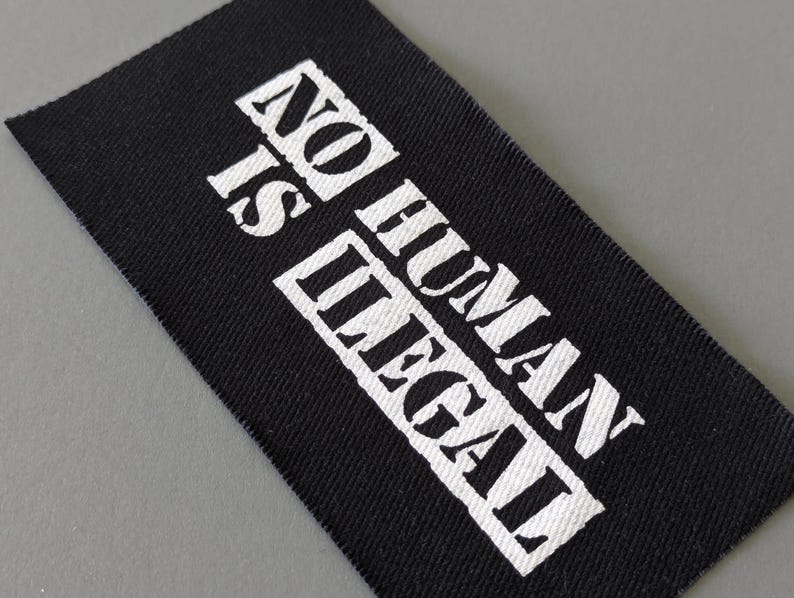 May include: Black rectangular fabric label with the text "NO HUMAN IS ILLEGAL" printed in white, bold, capital letters. The label is angled, with a gray background.