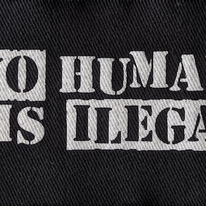 May include: Black fabric with the white text "NO HUMAN IS ILLEGAL" printed across the center. The text is in a bold, stencil-style font. The fabric has a textured appearance.