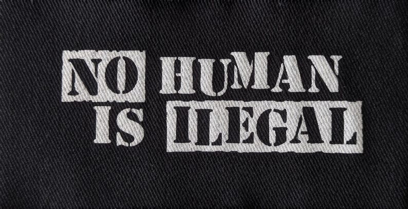 May include: Black fabric with the white text "NO HUMAN IS ILLEGAL" printed across the center. The text is in a bold, stencil-style font. The fabric has a textured appearance.