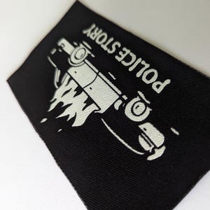 May include: Black fabric patch with white text that reads "POLICE STORY" and a white silhouette of a car on fire.