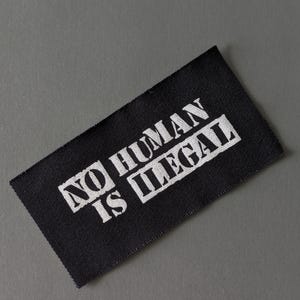 May include: A black rectangular fabric label with the words "NO HUMAN IS ILLEGAL" printed in white, set against a gray background. The text is in a bold, sans-serif font.
