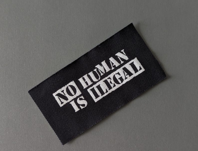 May include: A black rectangular fabric label with the words "NO HUMAN IS ILLEGAL" printed in white, set against a gray background. The text is in a bold, sans-serif font.