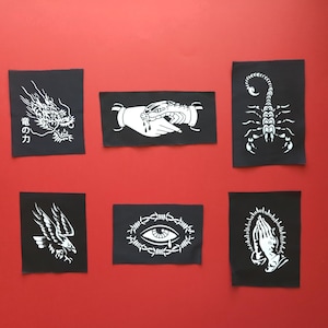 Puede incluir: Seis parches rectangulares de tela negra con diseños serigrafiados en blanco: un dragón con texto japonés, una mano sosteniendo una serpiente, un escorpión, un águila volando, un ojo rodeado de alambre de espino y manos rezando con halo.
