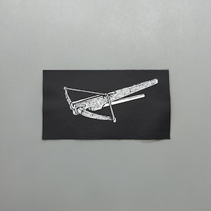 Op de afbeelding: Een zwarte rechthoekige patch met een witte illustratie van een kruisboog. Het kruisboogontwerp heeft gedetailleerde houtnerf- en snaarelementen. De patch staat tegen een grijze achtergrond.