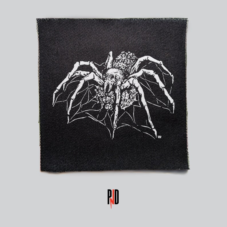 May include: A white spider with a large abdomen and many legs on a black fabric patch. The spider is in the center of the patch and is surrounded by a web. The patch is rectangular and has a rough texture.