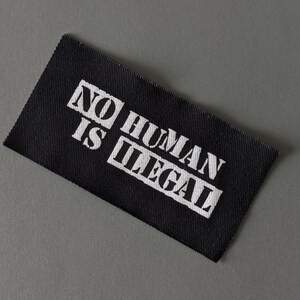 May include: Black rectangular fabric label with the text "NO HUMAN IS ILLEGAL" in white block letters. The label is angled and set against a gray background.