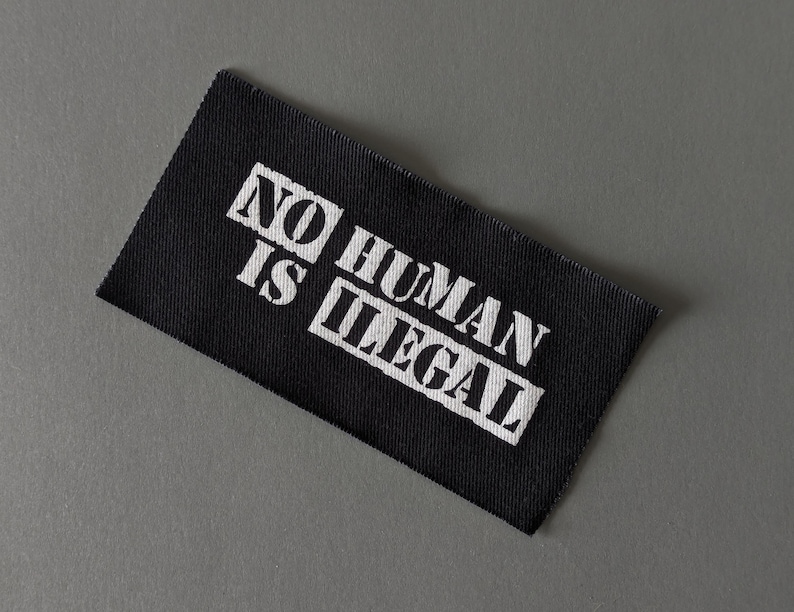 May include: Black rectangular fabric label with the text "NO HUMAN IS ILLEGAL" in white block letters. The label is angled and set against a gray background.