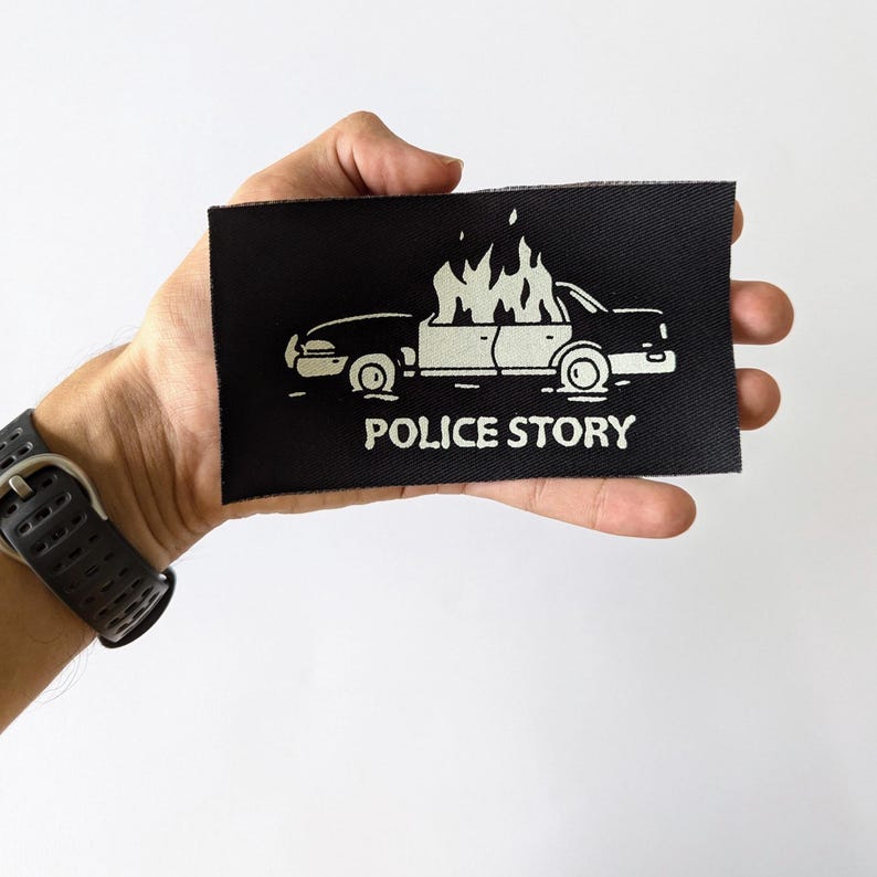 May include: Black fabric patch with a white outline of a car on fire and the text "POLICE STORY" underneath.