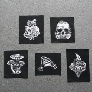 May include: Five black square patches with white illustrations. Designs include a skull with flowers, a mushroom with an eye, a skeleton hand, and a skull with a beard and flowers. The patches are arranged on a gray surface.