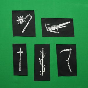 May include: Five black patches with white illustrations of medieval weaponry. The patches include a flail, crossbow, poleaxe, sword, and scythe. The patches are arranged on a green background.