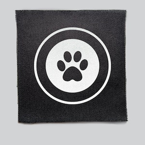 Pode incluir: Um patch quadrado de tecido preto com uma pegada branca dentro de um círculo branco. A pegada está centrada dentro do círculo, e o patch tem uma aparência texturizada. O design é simples e limpo.