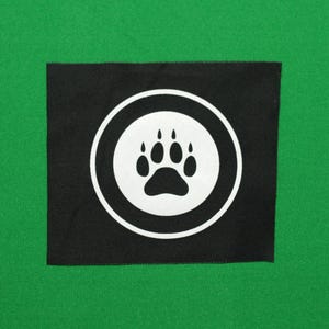 May include: A square, black patch with a white paw print inside a white circle. The paw print is centrally positioned within the circle, set against a green backdrop. This patch is suitable for pet owners.
