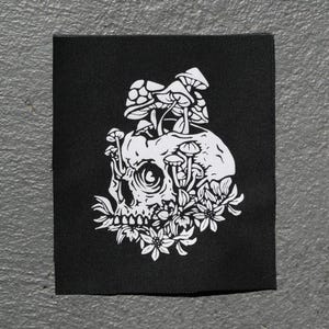 May include: Black square patch featuring a white illustration of a skull with mushrooms growing from it and flowers at the base. The design is detailed and gothic in style, suitable for clothing or accessories.