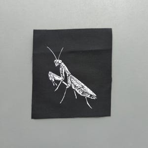 May include: A black square patch with a white illustration of a praying mantis. The insect is depicted in a detailed, stylized manner, with its legs and antennae clearly visible. The patch is likely for clothing or accessories.
