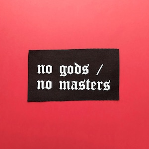 Puede incluir: Un parche rectangular de tela negra con texto bordado en blanco que dice no gods / no masters en una fuente de estilo gótico. El parche tiene una textura tejida visible y bordes limpios y rectos, presentando un diseño de alto contraste.