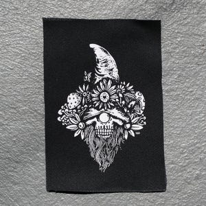 Könnte beinhalten: Schwarzer, rechteckiger Aufnäher mit einer weißen Illustration eines Schädels mit Zwergenhut, umgeben von Blumen und Pilzen. Der Aufnäher hat eine strukturierte Oberfläche.