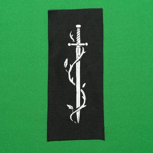 Puede incluir: Parche rectangular negro con un diseño de espada blanca entrelazada con vides. La espada tiene una empuñadura detallada y una hoja afilada. El parche está sobre un fondo verde.