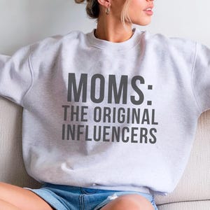 May include: A pale grey sweatshirt featuring the text "MOMS: THE ORIGINAL INFLUENCERS" in dark grey. The sweatshirt is worn by a person sitting on a white sofa. The person is wearing denim shorts.