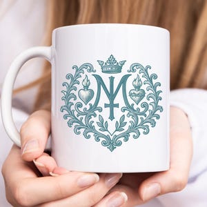 以下が含まれることがあります： 王冠、文字のM、2つのハート、花柄の要素が特徴の、ターコイズのデザインが施された白いセラミックマグ。 マグカップには湾曲したハンドルが付いています。