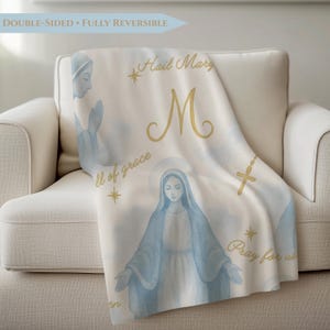 May include: A white, double-sided, fully reversible blanket draped over a white armchair. The blanket features a watercolor illustration of the Virgin Mary, gold text that reads "Hail Mary", "M", "full of grace", and "Pray for us", and a gold cross.
