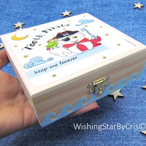 May include: A wooden tooth fairy box with a blue and white wave design around the edge. The lid features a cartoon tooth pirate with a parrot and the text "Tooth Pirate keep me forever".