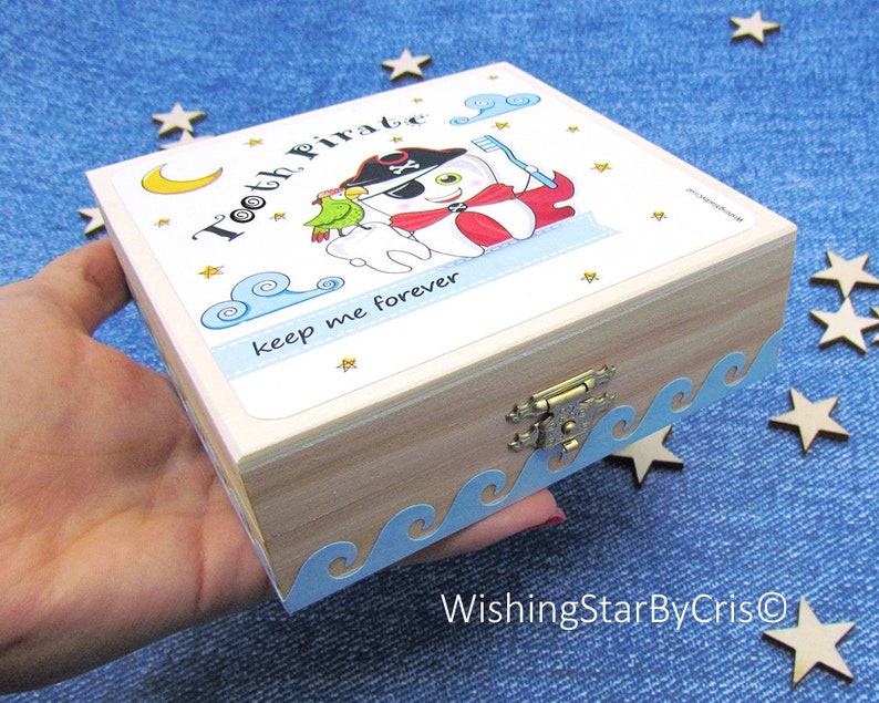 May include: A wooden tooth fairy box with a blue and white wave design around the edge. The lid features a cartoon tooth pirate with a parrot and the text "Tooth Pirate keep me forever".