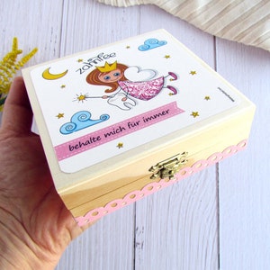 May include: A wooden box with a pink and white lid featuring a cartoon tooth fairy with a tooth and a wand. The lid has the text "Zahnfee" and "behalte mich f&uuml;r immer".