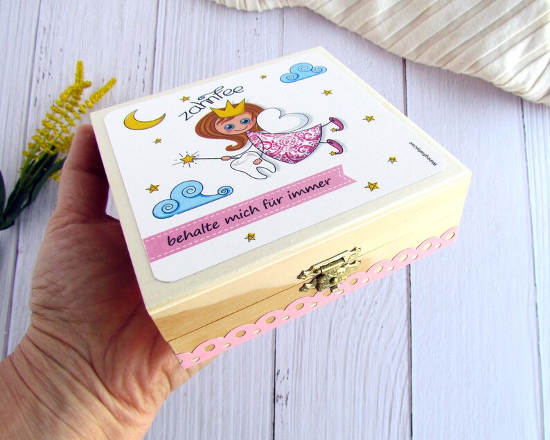 May include: A wooden box with a pink and white lid featuring a cartoon tooth fairy with a tooth and a wand. The lid has the text "Zahnfee" and "behalte mich f&uuml;r immer".