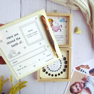 May include: A wooden box with a white background and a small fairy illustration. The box has a compartment for storing baby teeth and a space for a photo. The text on the box reads "Here are stored the teeth of..." and includes a space for the child's name and birth date.