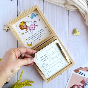 May include: A wooden box with a hinged lid, decorated with a whimsical illustration of a tooth fairy. The box is open, revealing a white card with the text "Here are stored the teeth of" and a space for a photo and other details. The box is a tooth fairy keepsake box for storing baby teeth.