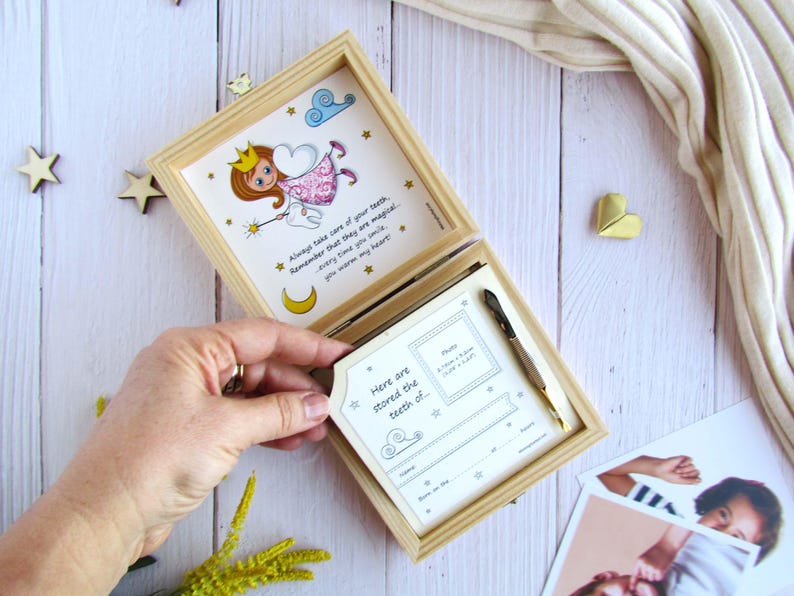 May include: A wooden box with a hinged lid, decorated with a whimsical illustration of a tooth fairy. The box is open, revealing a white card with the text "Here are stored the teeth of" and a space for a photo and other details. The box is a tooth fairy keepsake box for storing baby teeth.