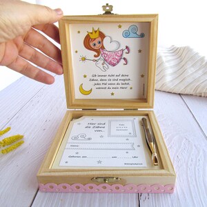 May include: A wooden box with a pink and white design. The box is open and has a small card inside that says "Here are the teeth from..." The box also has a small illustration of a tooth fairy with a wand and a crown. The text on the box says "Give always attention to your teeth, because they are magic. Every time you laugh, you warm my heart."
