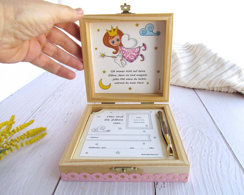 May include: A wooden box with a pink and white design. The box is open and has a small card inside that says "Here are the teeth from..." The box also has a small illustration of a tooth fairy with a wand and a crown. The text on the box says "Give always attention to your teeth, because they are magic. Every time you laugh, you warm my heart."