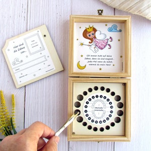 May include: Wooden box with a tooth fairy theme. The box has a compartment for storing baby teeth and a chart to track lost teeth. The box is decorated with a tooth fairy illustration and the text "Gib immer Acht auf deine Z&auml;hne, denn sie sind magisch. Jedes Mal wenn du lachst, w&auml;rmst du mein Herz!"