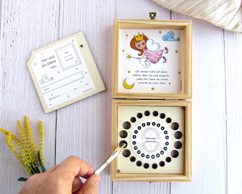 May include: Wooden box with a tooth fairy theme. The box has a compartment for storing baby teeth and a chart to track lost teeth. The box is decorated with a tooth fairy illustration and the text "Gib immer Acht auf deine Z&auml;hne, denn sie sind magisch. Jedes Mal wenn du lachst, w&auml;rmst du mein Herz!"