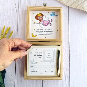 May include: A wooden box with a hinged lid. The inside of the box has a compartment for a photo and a small space for a tooth. The box is decorated with a cartoon tooth fairy and the text "Gib immer Acht auf deine Zahne, denn sie sind magisch. Jedes Mal wenn du lachst, warmst du mein Herz!"