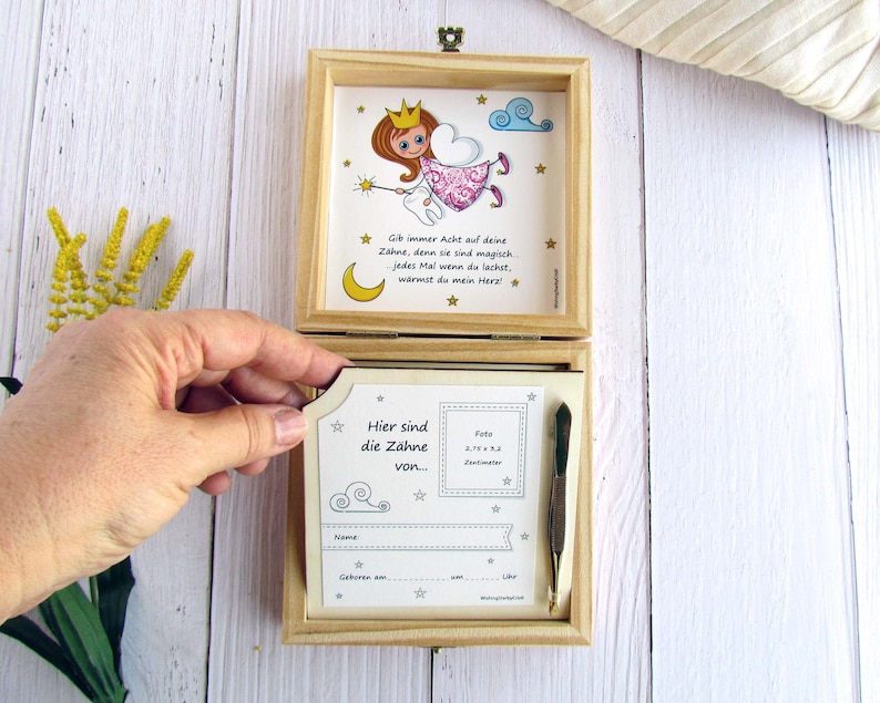 May include: A wooden box with a hinged lid. The inside of the box has a compartment for a photo and a small space for a tooth. The box is decorated with a cartoon tooth fairy and the text "Gib immer Acht auf deine Zahne, denn sie sind magisch. Jedes Mal wenn du lachst, warmst du mein Herz!"