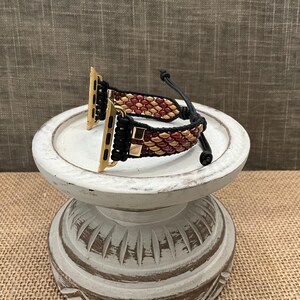 May include: A black and gold beaded Apple Watch band with a red and gold beaded design. The band is shown on a white pedestal.