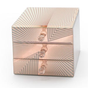 May include: A square mirrored jewelry box with three stacked drawers. Each drawer front features a rose gold geometric sunburst pattern and a clear, faceted crystal-style knob. The reflective surfaces create a repeating linear design.