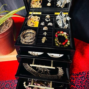 May include: A black multi-tiered jewelry box with a hinged lid and three pull-out drawers. The interior is lined with black velvet, containing various rings, bracelets, beaded necklaces, and metallic ornaments organized in dedicated compartments.