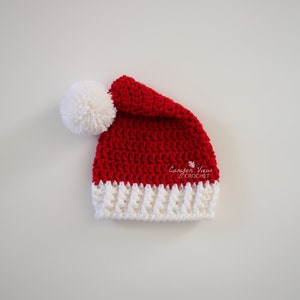 Puede incluir: Un gorro de Pap&aacute; Noel de ganchillo hecho a mano, rojo y blanco, con un pomp&oacute;n blanco. El gorro presenta un borde blanco acanalado y est&aacute; hecho con un punto texturizado. El logotipo de Canyon View Crochet es visible.