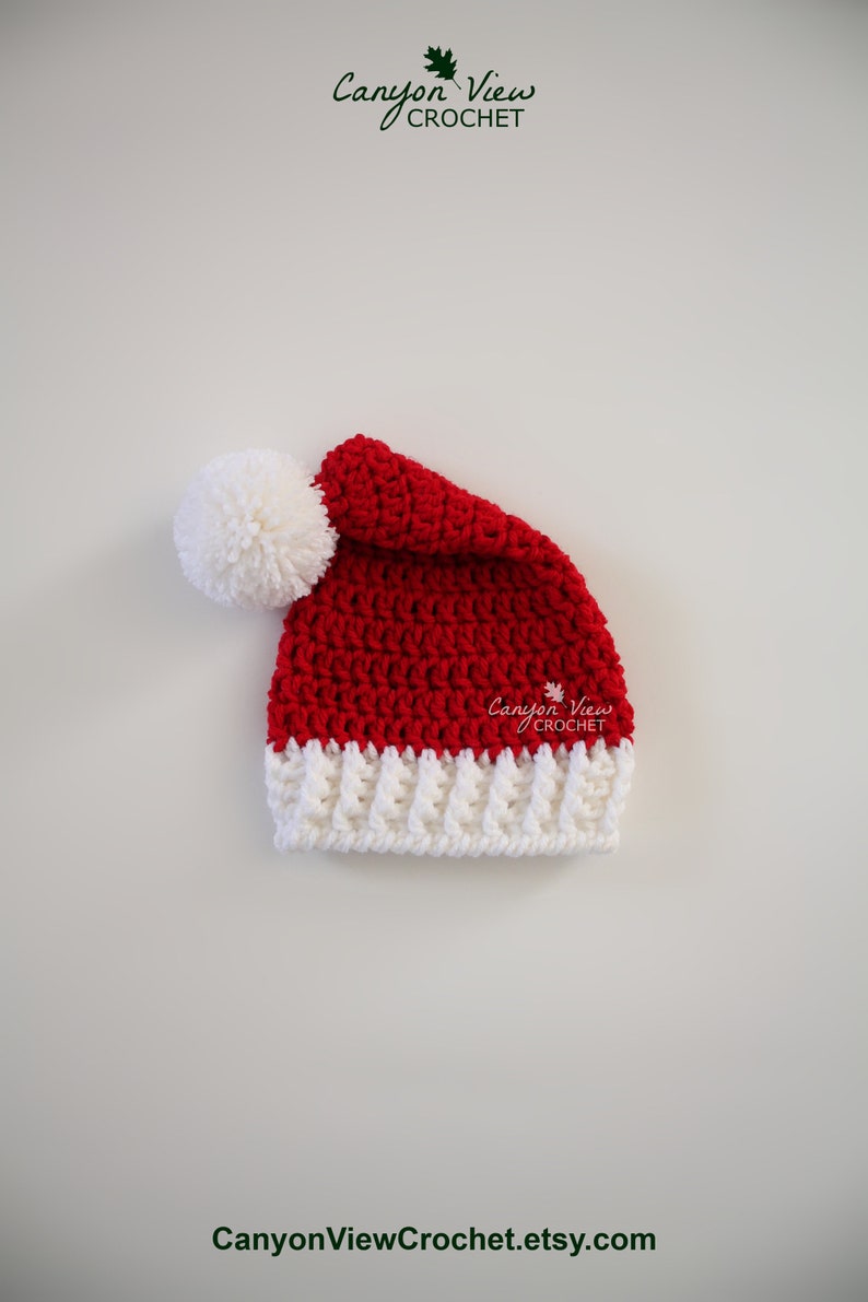 Puede incluir: Un gorro de Pap&aacute; Noel de ganchillo hecho a mano, rojo y blanco, con un pomp&oacute;n blanco. El gorro presenta un borde blanco acanalado y est&aacute; hecho con un punto texturizado. El logotipo de Canyon View Crochet es visible.