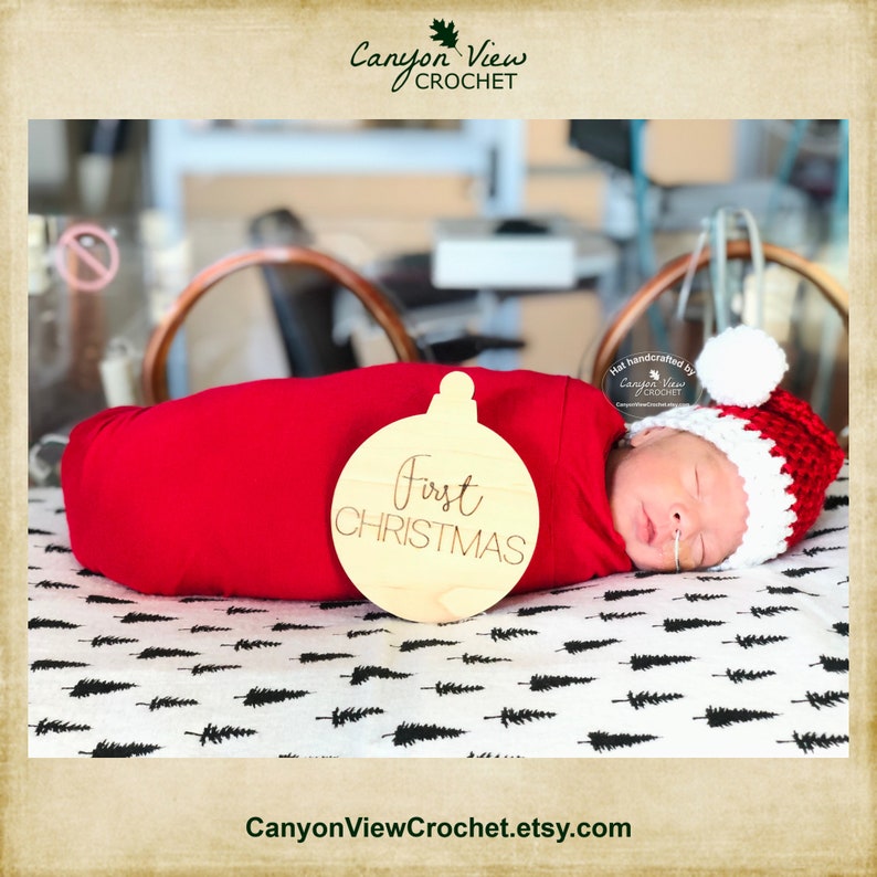 May include: A newborn baby wearing a red and white crocheted Santa hat is swaddled in a red blanket. A wooden ornament with the words "First Christmas" is in front of the baby.