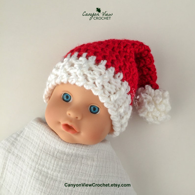 May include: A red and white crocheted Santa hat with a white pom-pom on the end. The hat is on a baby doll's head.