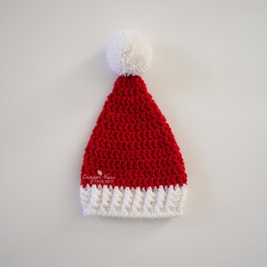 Puede incluir: Un gorro de Pap&aacute; Noel rojo y blanco tejido a ganchillo, con un pomp&oacute;n blanco. El gorro tiene un borde blanco acanalado y est&aacute; hecho de hilo texturizado. El texto "Canyon View Crochet" y "CanyonViewCrochet.etsy.com" son visibles.