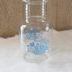 May include: Clear glass pitcher with a flared spout and a ribbed neck. The pitcher features a light blue floral design and measurement markings. The text at the bottom reads "NOT USE METAL STIRRER".