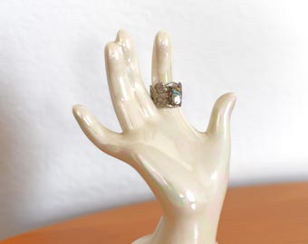 Op de afbeelding: Een parelmoer keramische ringhouder in de vorm van een hand met een zilverkleurige ring. De ring heeft een sierlijk ontwerp met een centrale steen. De ringhouder wordt op een houten oppervlak gepresenteerd.